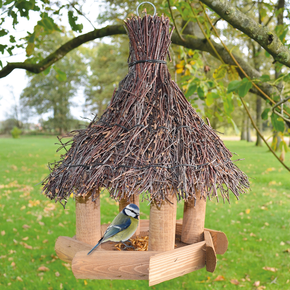 Vogelhaus Sweet Home mit einem Dach aus Reisig und einem stabilen Holzrahmen, an dem eine Blaumeise auf der unteren Plattform sitzt