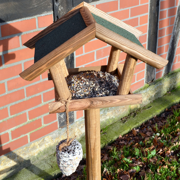 Vogelhaus Bird House aus Holz mit Futterspender an der Seite vor einer Backsteinwand und auf einem Rasenbereich aufgestellt