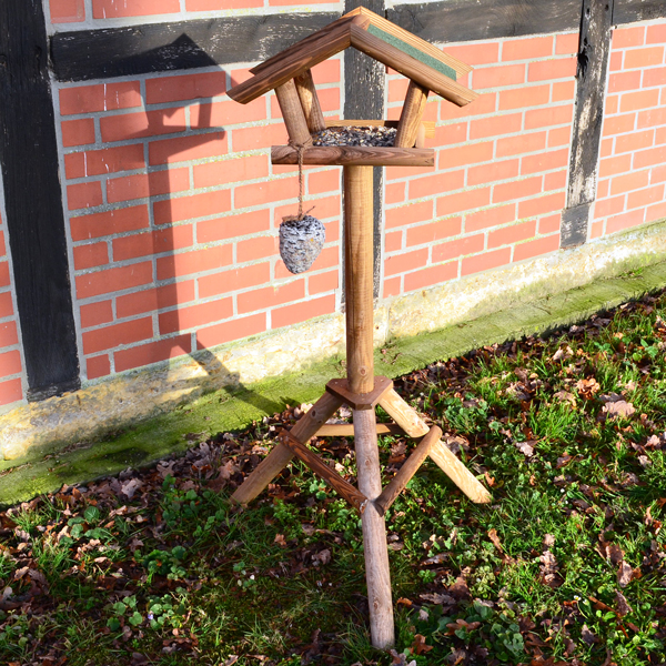 Vogelhaus Bird House auf einem stabilen Holzständer aufgestellt im Freien vor einer Hauswand mit Futterspender an der Seite Das Vogelhaus steht auf einem mit Gras und Blättern bedeckten Boden