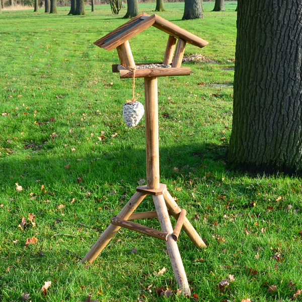 Vogelhaus Bird House auf einem stabilen Holzständer in einem offenen grünen Rasenbereich aufgestellt neben einem Baum mit einem Futterspender an der Seite des Hauses