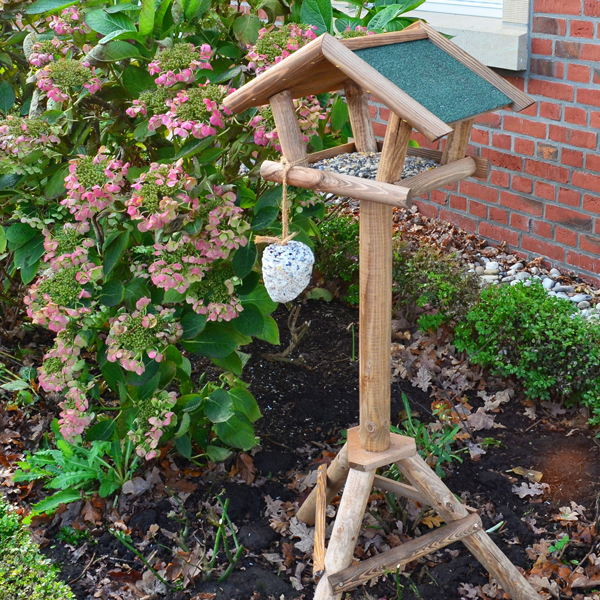 Vogelhaus Bird House mit robustem Ständer, platziert in einem gepflegten Garten, umgeben von grünen Pflanzen und Blüten. Eine Futterstation mit Futterspender für Vögel hängt an der Seite des Vogelhauses